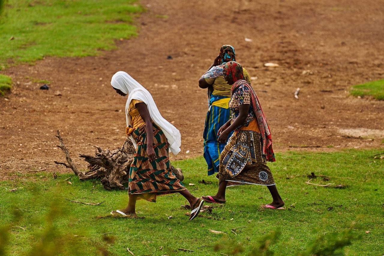 Three women stroll through a grassy area in Minna, Nigeria, wearing colorful traditional clothing.